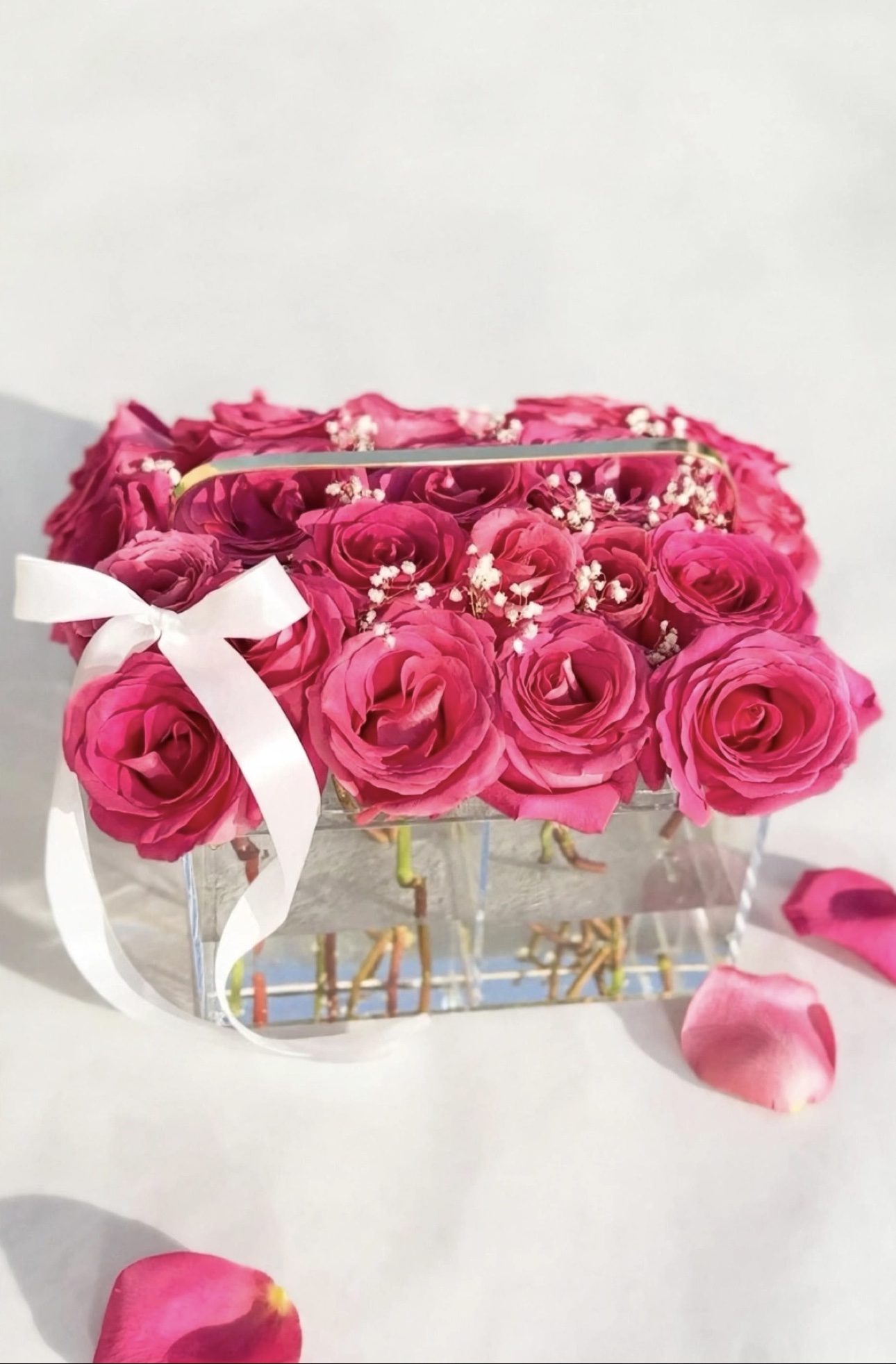 Pink roses with baby's breath in a clear acrylic box with a gold handle, tied with a white ribbon bow, with rose petals scattered beside it